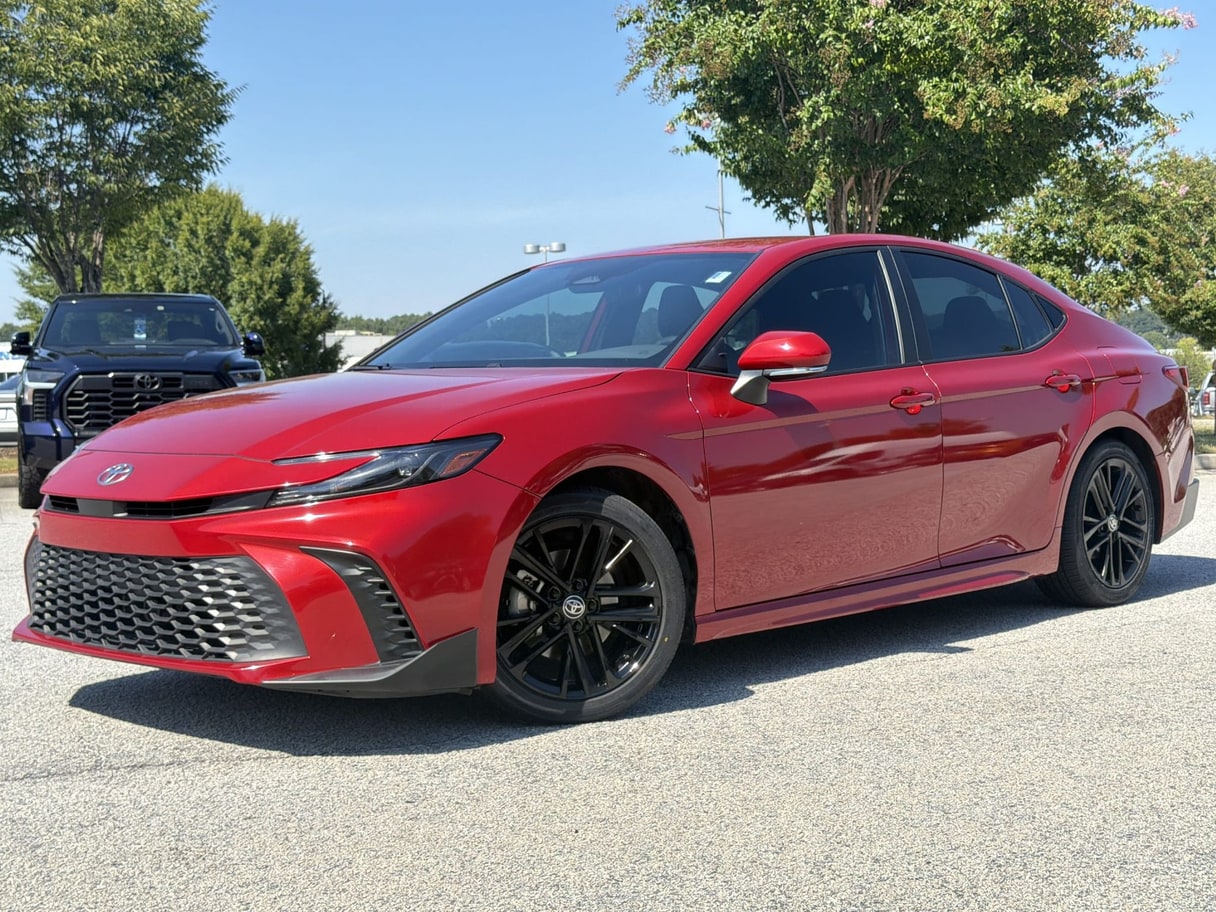 A red 2025 Toyota Camry sedan on a dealership lot with trees and other vehicles behind it.
