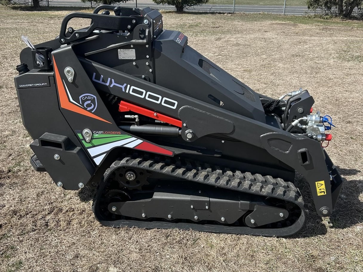 A black CAST WX1000 tracked mini skid steer on grass beside a fence and trees.