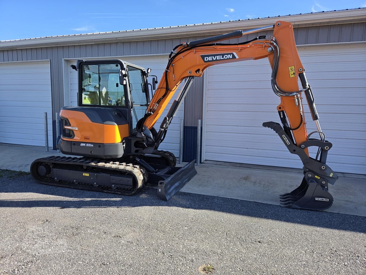 An orange Develon DX55R-7 mini excavator in front of a grey metal building.