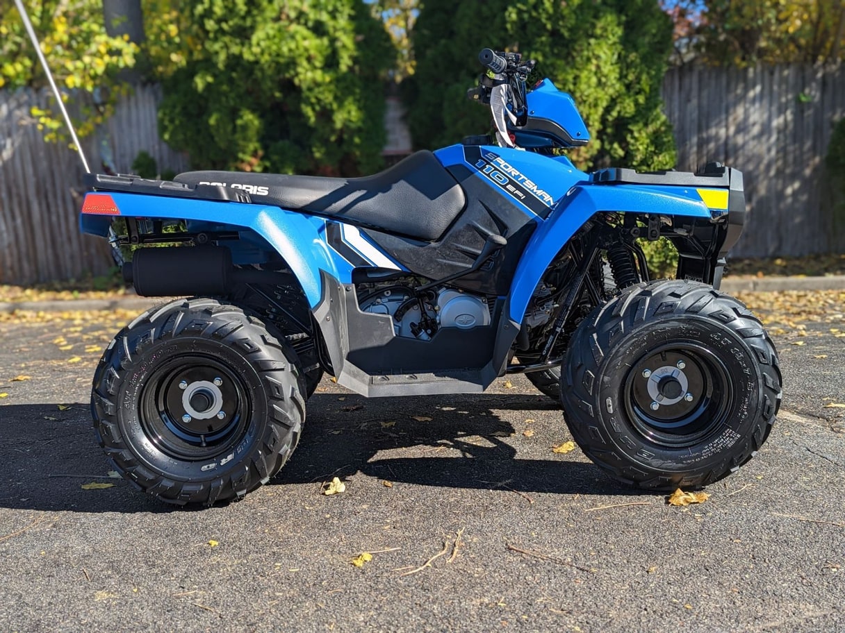 A blue Polaris Sportsman 110 ATV on an asphalt lot in front of a wooden fence.