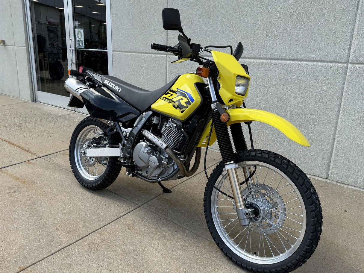 A yellow 2027 Suzuki DR650S dirt bike on pavement in front of a dealership wall.