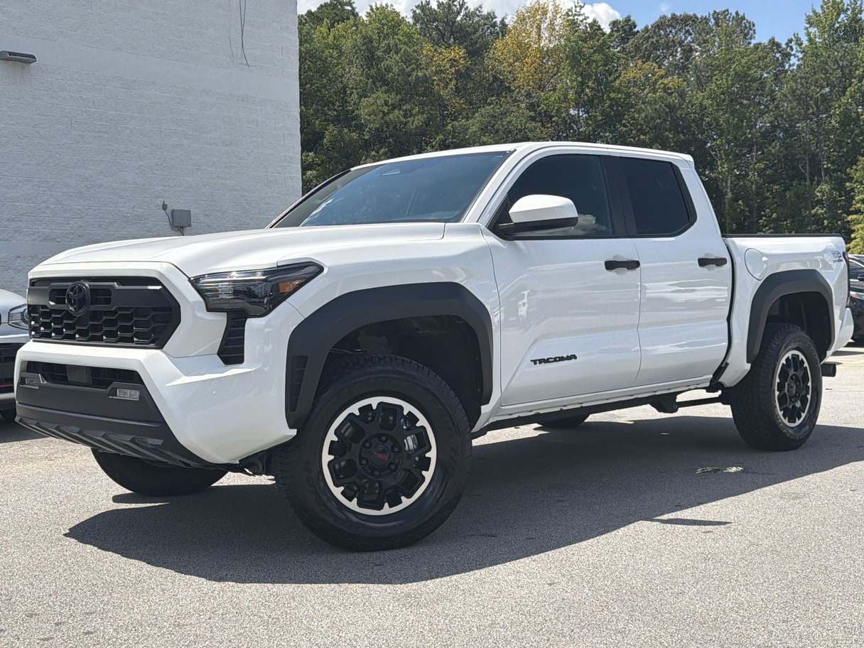 A white 2026 Toyota Tacoma crew-cab pickup on a dealership lot, with a wall and trees behind it.